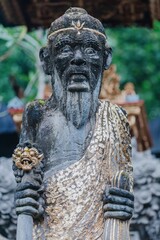 A weathered stone statue of a Balinese Hindu deity stands in Ubud, Bali, Indonesia. The figure is adorned with gold leaf and holds a symbolic object.