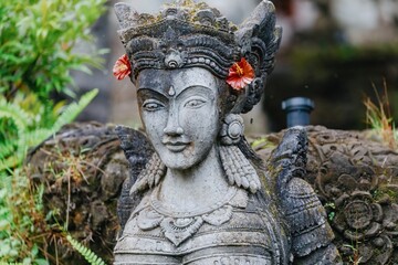 A weathered stone statue of a Balinese deity stands guard in a lush garden in Ubud, Bali, Indonesia. The statue is adorned with flowers, representing offerings and reverence.