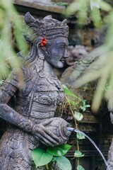 Stone statue of Balinese woman with flower in hair, holding vessel with water pouring out, in Ubud, Bali, Indonesia. The statue is part of a water feature.