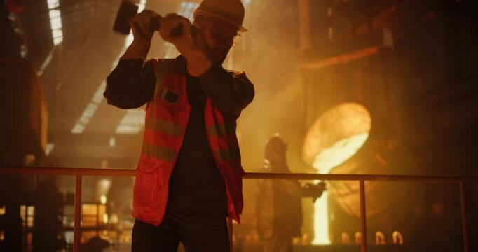 Young Man Using Brute Force, Swinging Sledgehammer in an Iron Ore Foundry. Operators Transporting Liquid Metal From an Industrial Furnace and Pouring It Into a Mold in the Background