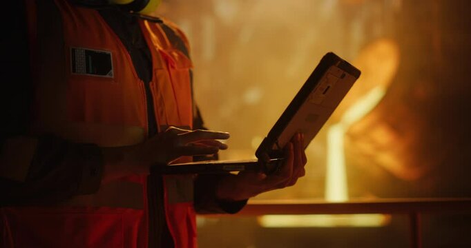 Close Up of Engineer Using Laptop Computer while Hot Metal Pouring Down in a Furnace in the Background. Specialist Working on Modern Monitoring Technology in a Heavy Industry Foundry