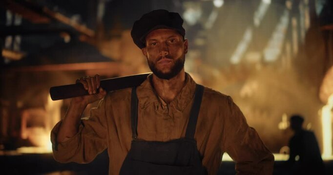 Working-Class Man of the Industrial Era Posing in Front of Camera While Carrying a Sledgehammer Over His Shoulder. Labor Worker Walking From an Iron Foundry in the Background