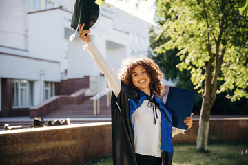 Achievement in life, with diploma. Cheerful graduate student in a gown is standing outdoors