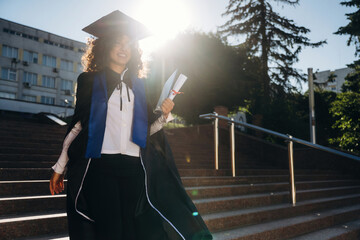 Stairs and university behind. Cheerful graduate student in a gown is standing outdoors