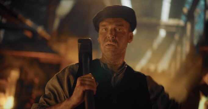 Foundryman of the Industrial Era Posing in Front of the Camera While Placing a Heavy Sledgehammer Over His Shoulder. Labor Worker Standing with an Iron Foundry in the Background