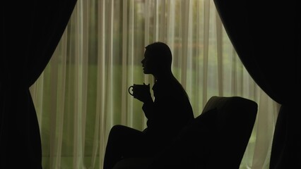 Silhouette of a female, distressed sitting on a sofa and holding a cup of hot tea.