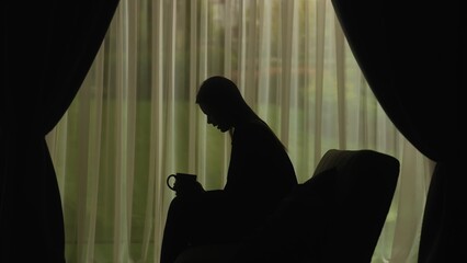 Silhouette of a female, distressed sitting on a sofa and holding a cup of hot tea.