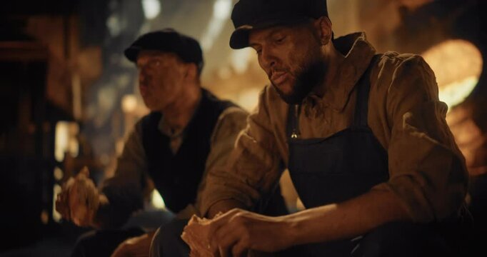 Worn-Out Male Factory Workers of the Industrial Revolution Pause For a Lunch Break to Eat a Loaf of Bread by the Furnace. Their Clothing is heavily Covered in Soot, Expressions on Faces Shows Fatigue
