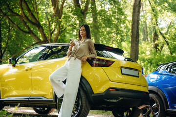 Leaning on the vehicle, using phone. Young woman is in the forest with two cars