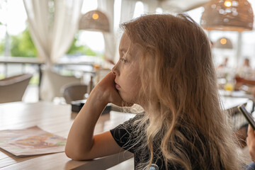  Young girl gazing thoughtfully, seated in a well-lit café. Her right hand rests on her chin, and...