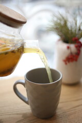 Pouring hot herbal tea into a ceramic mug from a glass teapot on a wooden table. Cozy warm drink in natural daylight. Winter or autumn relaxation mood, healthy lifestyle concept.
