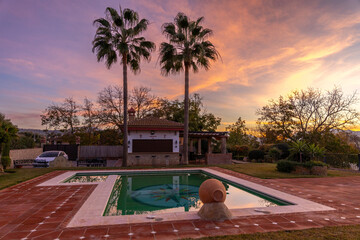 Golden hour at a countryside house with a colorful sky of orange, purple, and blue hues, palm trees in the background, and a pool in the foreground. A serene and vibrant scene.