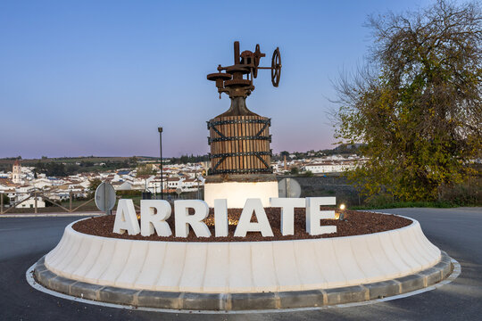Entrance sign to Arriate village with the town visible in the background under a blue hour sky featuring purple and blue hues. A calm and charming Andalusian scene.