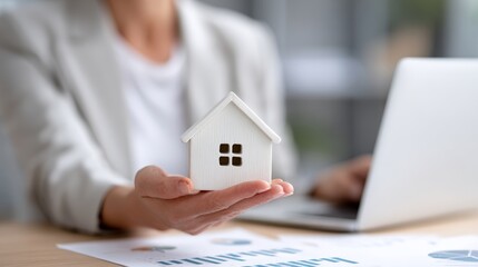 Real estate professional presenting miniature house model near laptop, displaying property market charts during client consultation for home buying services