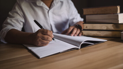 Person writing in open book with pen at wooden desk. Student studying, author creating, researcher...
