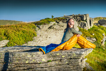 Young woman sitting in sleeping bag on a mountain cliff, enjoying the morning sun with eyes closed
