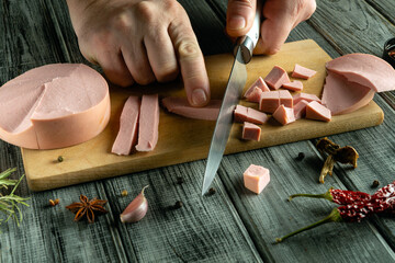 A person skillfully slices deli meats into cubes and strips on a wooden cutting board. Colorful spices and herbs are spread around, enhancing the culinary atmosphere