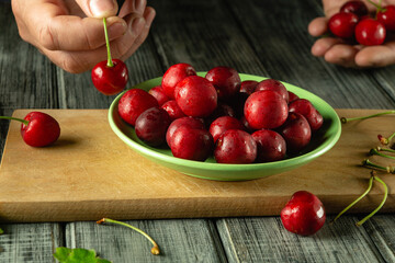 Hands carefully select ripe cherries from a wooden surface to add to a green bowl filled with glistening, fresh fruit in natural light