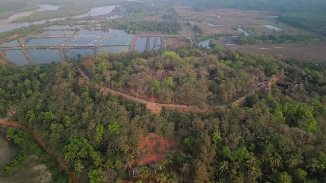 Remains of Chapora Fort nestled in Goa's dense forest. Red paths lead to the ruins with round bastions. Nearby rivers and fields reflect early morning light