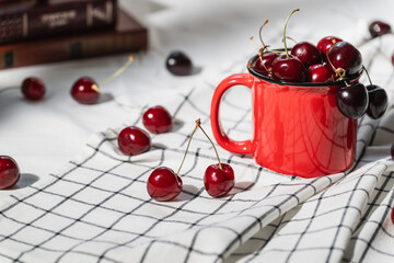 A ripe red cherry in a red mug and a stack of books on a checkered napkin close-up. Selective focus.
