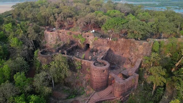 Remains of Chapora Fort nestled in Goa's dense forest. Colonial-era fort entrance with round towers above forested slope. High coastal cliffs meet green water and white sands in Goa, India