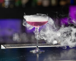 This visually striking pink alcoholic cocktail in a coupe glass features a large with more smoke...
