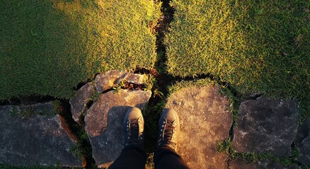Person Standing on Cracked Stone Walkway Over Grass During Sunset, Long Shadows Cast on Broken Surface and Green Lawn Below