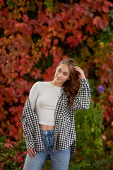 Young woman in casual autumn outfit posing against vibrant red ivy wall, stylish fashion portrait...