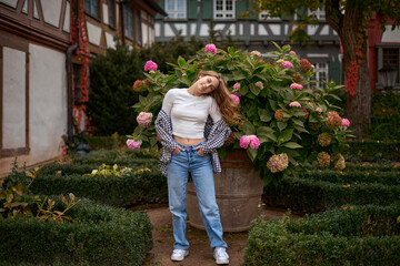 Beautiful Young Woman Flipping Hair in a Lush Garden with Pink Hydrangeas and Traditional European Houses. Young woman posing with hydrangeas in a European garden, surrounded by half-timbered houses