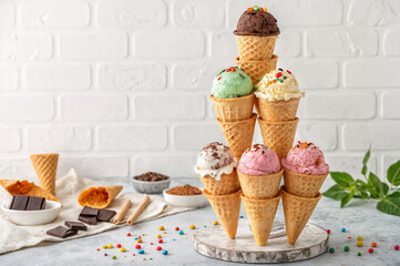 shows a pyramid of ice cream cones with various flavors, including chocolate, green, yellow, and pink, arranged on a wooden stand. The background features a light-colored brick wall and a few scattere