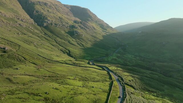 Car moving up sunrise illuminated mountain pass with camera pan up showing mountaintops. Summer. Kirkstone Pass, Lake District, Cumbria, UK.