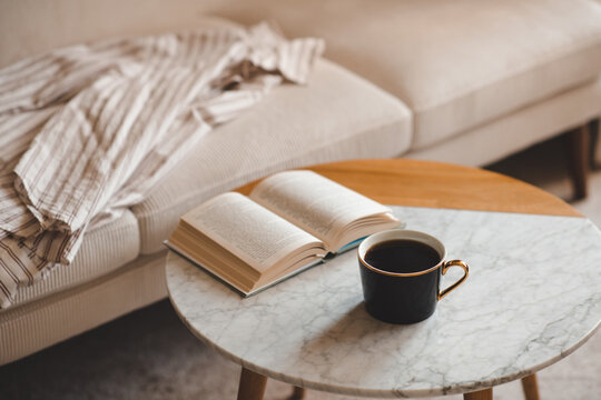 Cup of black fresh coffee with open paper book staying on marble table and velvet couch in living room close up. Evening time. Relaxation.