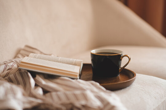 Cup of black tea with open paper book and pajamas shirt cloth on wooden tray in couch in living room close up. Good morning. Breakfast time.