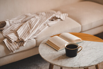 Cup of black fresh coffee with open paper book staying on marble table and velvet couch in living room close up. Evening time. Relaxation.