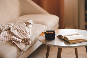 Mug of fresh black tea with open paper book staying on marble table and velour couch in living room close up. Night time. Relaxation.