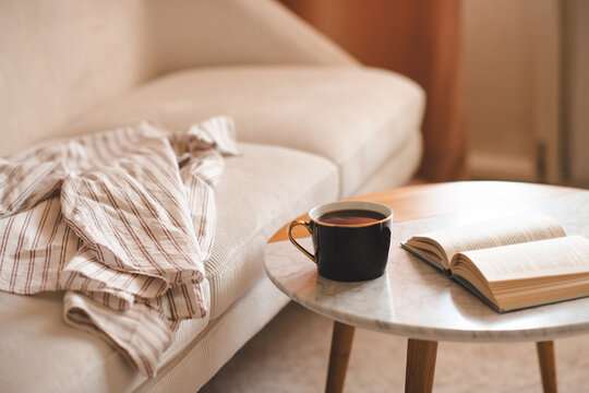 Cup of black fresh coffee with open paper book staying on marble table and velvet couch in living room close up. Evening time. Relaxation.