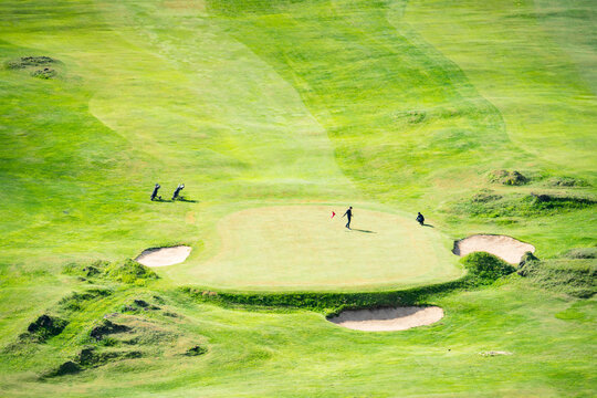 View of golfers enjoying a sunny day on a lush green course with sand traps and a red flag waving gently in the breeze, VestmannaeyjabÃ¦r, VestmannaeyjabÃ¦r, Iceland.
