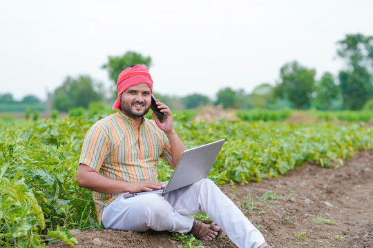 young indian farmer sitting at green agricultural field using laptop talking on smartphone