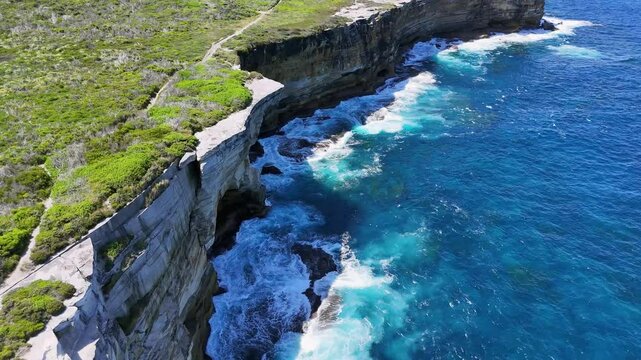 Sydney, Australia -UHD Drone Video- Spectacular view from above of waves splashing on the sandstone cliffs of the coast, of the scenic Cape Baily Track and Lighthouse in Kamay National Park.