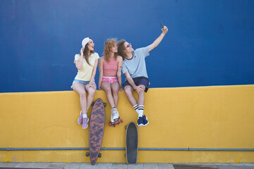 Group of trendy friends taking a selfie while sitting on a colorful wall with skateboards