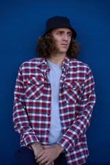 Young man wearing bucket hat and checkered shirt leaning against blue wall