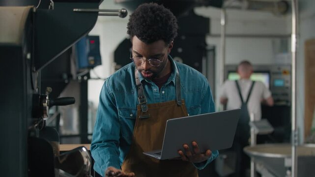 African American ethnic male guy man expert in coffee industry check fresh roasted beans typing laptop online going away inspector inspect roasting machine quality roast with computer small business