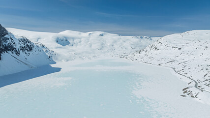 Aerial view of a serene, icy lake nestled between snow-covered mountains under a clear blue sky, creating a stark contrast of textures and tones, Geiranger, MÃ¸re og Romsdal, Norway.