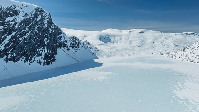 Aerial view of a frozen lake nestled between snow-capped mountains, contrasting the stark white against the clear blue sky, Geiranger, M&Atilde;&cedil;re og Romsdal, Norway.