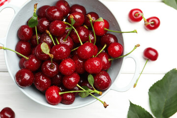 Fresh wet cherries in colander and leaves on white wooden table, flat lay
