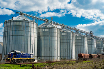 A train sits beside several large silos in a storage facility under a partly cloudy sky,...