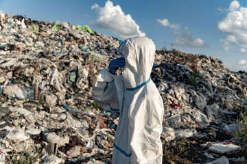 A person in protective clothing observes a landfill overflowing with plastic and debris on a clear day, highlighting the pollution crisis