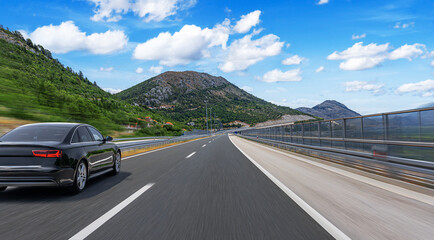 Black car driving on a modern mountain highway with a clear blue sky, surrounded by green hills and...