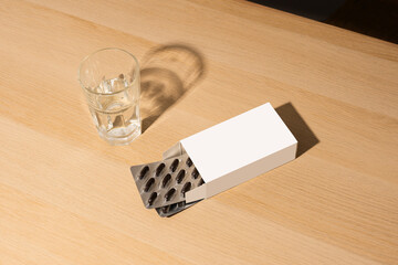 mockup scene with a white blank box, visible blister packs, and a glass of water on rustic wood....
