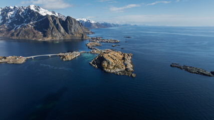 Aerial view of Reine's rugged coastline meets the deep blue sea, a bridge connecting islands beneath snow-capped mountains, Reine, Nordland, Norway.